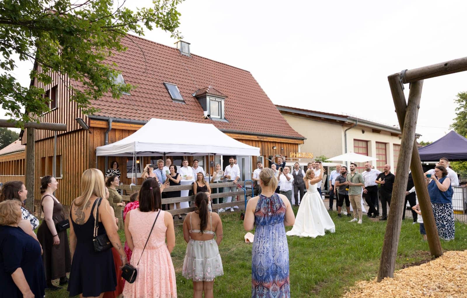 Außenansicht der Eventlocation EvenTraum Lola in Marxzell mit einer Hochzeitsgesellschaft im Hof, die gespannt den Wurf des Brautstraußes durch die Braut beobachtet.